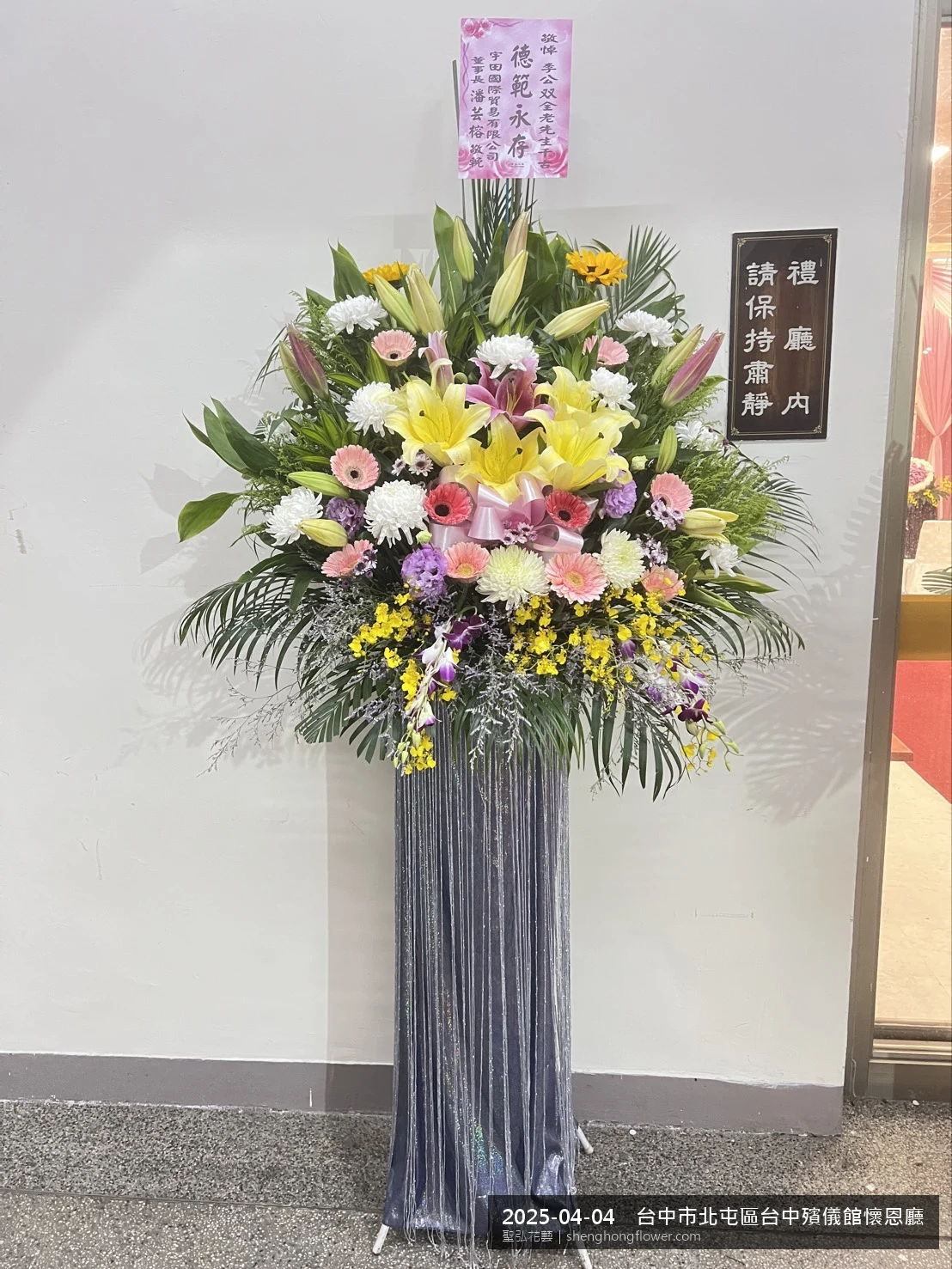 台中北屯台中殯儀館懷恩廳前置高架白菊花籃，氛圍莊嚴端莊。挑高花架穩固、字牌方向清晰，於春末穀雨時節氣候溫暖偏濕就位完成，陪伴家屬度過難捨時刻，聖弘花藝在地團隊現場實拍照。