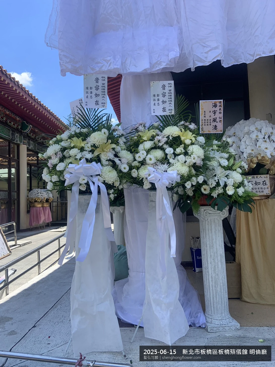 盛夏午後氣候炎熱下，新北板橋板橋殯儀館 明倫廳前光線柔和。我們選用高架白菊花籃，雙劍蘭挑高、黃菊鋪底，現場顯得陳設整齊，依指定方位整齊就位，聖弘花藝在地團隊現場實拍照。