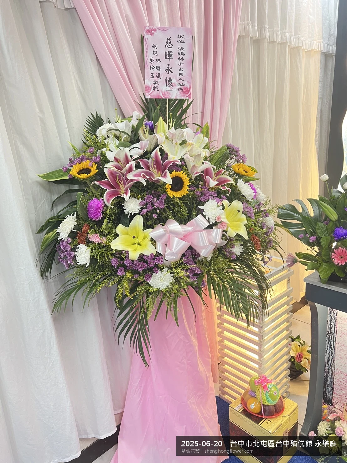 台中北屯台中殯儀館 永樂廳前置高架白菊花籃，氛圍素雅莊重。挑高花架穩固、字牌方向清晰，於盛夏午後氣候炎熱就位完成，陪伴家屬度過難捨時刻，聖弘花藝在地團隊現場實拍照。