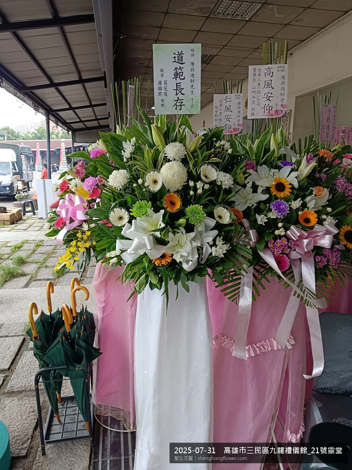 高雄三民九龍禮儀館_靈堂現場置高架白菊花籃，氛圍陳設整齊。挑高花架穩固、字牌方向清晰，於盛夏陽光氣候悶熱就位完成，於現場完成定位，聖弘花藝在地團隊現場實拍照提供在地專業配送服務。